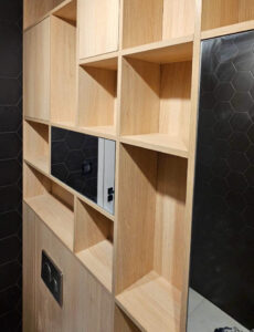 Zabudowa łazienki z płyty w kolorze jasnego drewna, z płytkami heksagon i zabudową geberit toalety. Lustra wkomponowane w ściankę z półkami i oświetlenie LED.