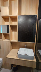 Zabudowa łazienki z płyty w kolorze jasnego drewna, z płytkami heksagon i zabudową geberit toalety. Lustra wkomponowane w ściankę z półkami i oświetlenie LED.