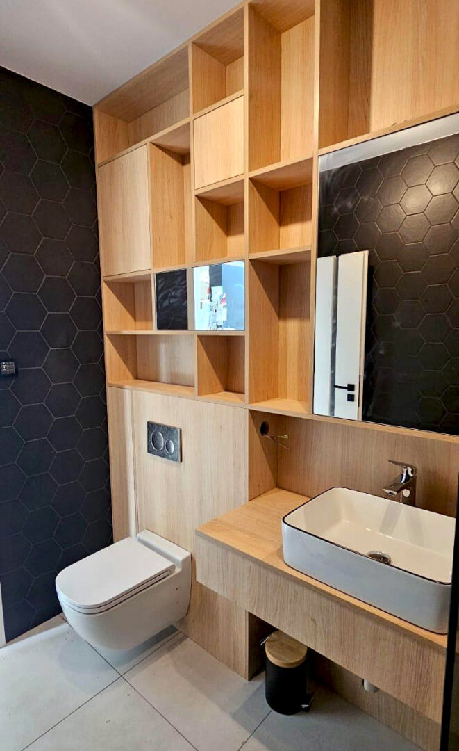 Zabudowa łazienki z płyty w kolorze jasnego drewna, z płytkami heksagon i zabudową geberit toalety. Lustra wkomponowane w ściankę z półkami i oświetlenie LED.